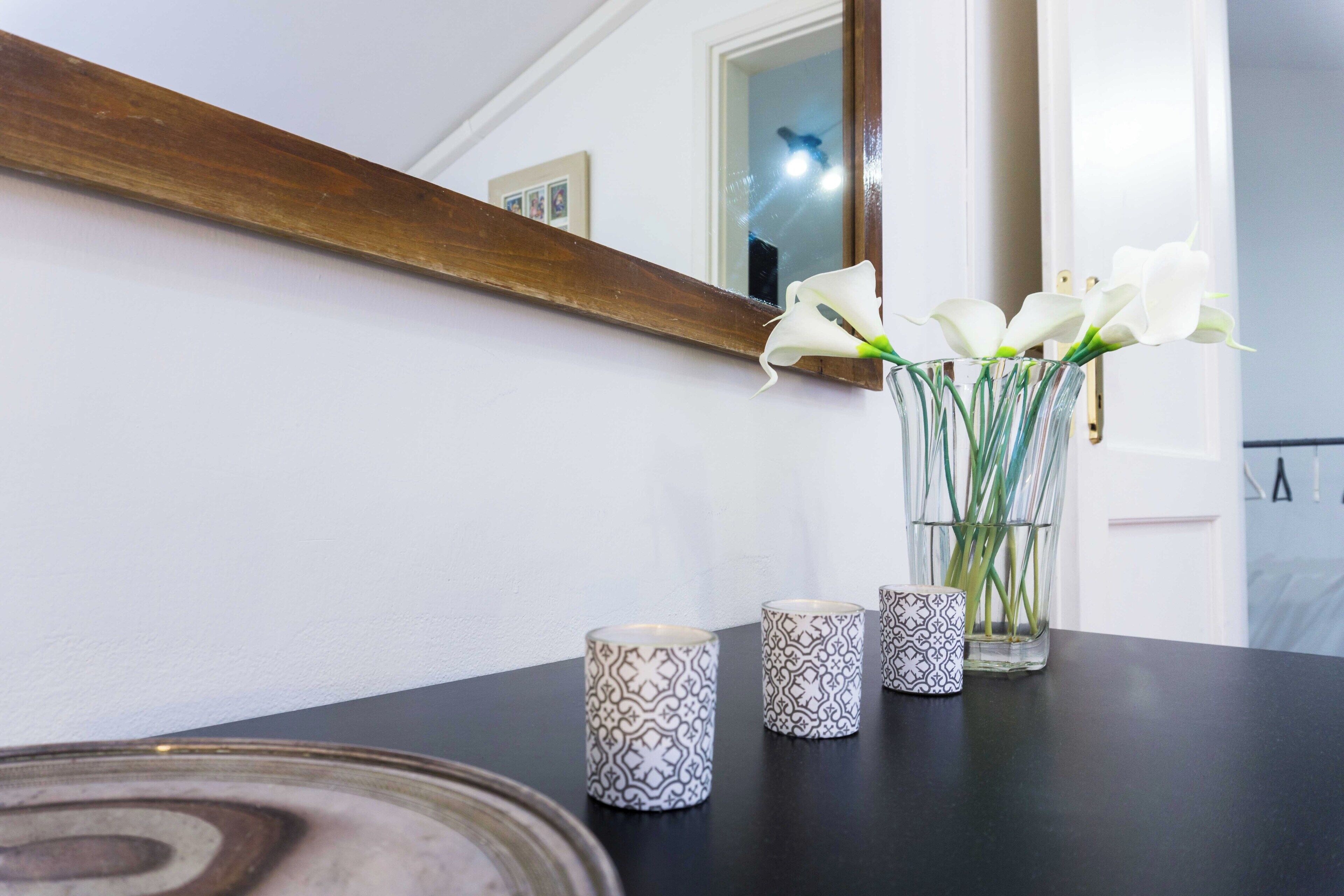 A table with a vase of white flowers, two patterned candles, and a mirror reflecting a door and a light fixture.