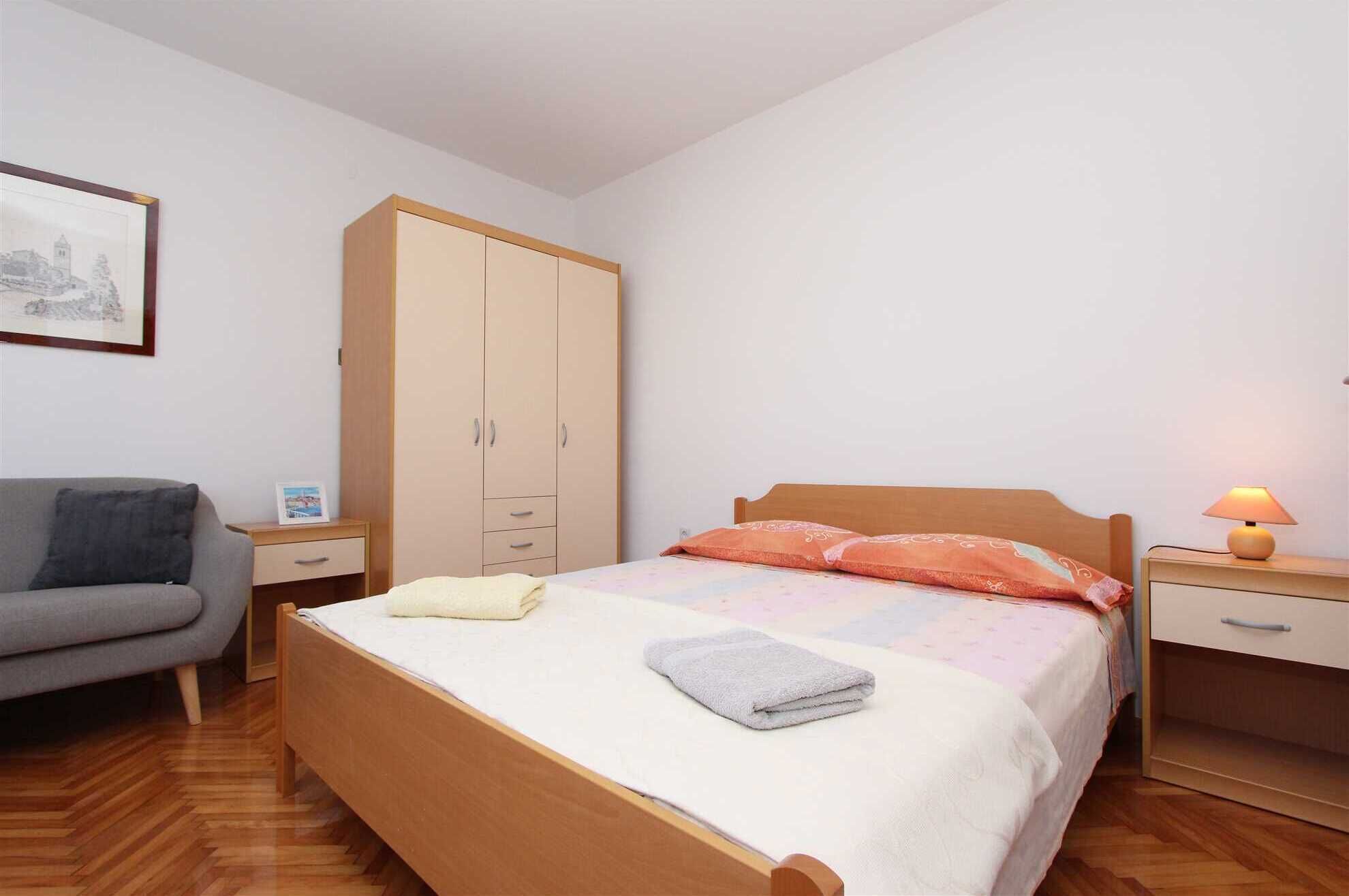 A bedroom with a bed, a wardrobe, a nightstand, a lamp, and a chair.