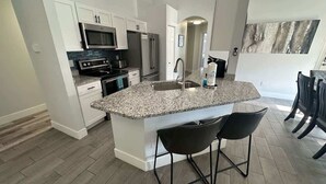 A modern kitchen with white cabinetry, a granite countertop, stainless steel appliances, and a bar area with two stools. - Signature Eight Home (Haines City)