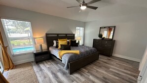 A bedroom with a bed, nightstands, a dresser, a ceiling fan, and a window with blinds. - Signature Eight Home (Haines City)
