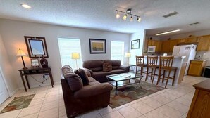 A living room with a sofa, coffee table, and dining area with chairs. There are framed pictures on the walls, a lamp, and a kitchen area with cabinets and appliances. - Sunset Dunes Villa (Haines City)