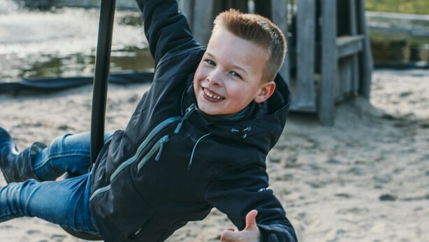 Un enfant sur une balançoire dans un terrain de jeux, avec une structure en bois et un bâtiment en arrière-plan.
