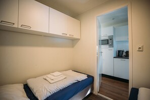 A small, compact room with a single bed, a kitchenette with a microwave, and a door leading to another room.
