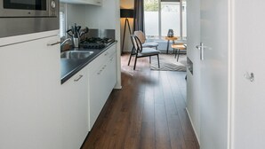 Un couloir étroit, avec une cuisine d’un côté, comprenant un four à micro-ondes et des placards. Le sol est en bois, et on aperçoit au loin un coin repas avec une table et des chaises.