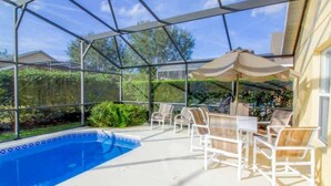 Un área de piscina exterior techada con sillones y sombrillas, rodeada de vegetación.
