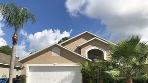 A single-story house with a beige exterior, a white garage door, and a small front yard with palm trees. - The Palms at Southern Dunes (Haines City)