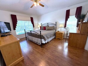 Un dormitorio con cama, mesitas de noche, escritorio, ventilador de techo y ventanas con cortinas.