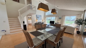 Un vaste espace de vie comprenant une table à manger avec un plateau en verre, des chaises en osier, un escalier, un canapé et une grande fenêtre offrant une vue dégagée.