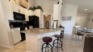 A kitchen with white cabinets, a granite countertop, a black refrigerator, and a microwave. There are two stools and a dining table with chairs. - Rebecca’s Villa (Haines City)