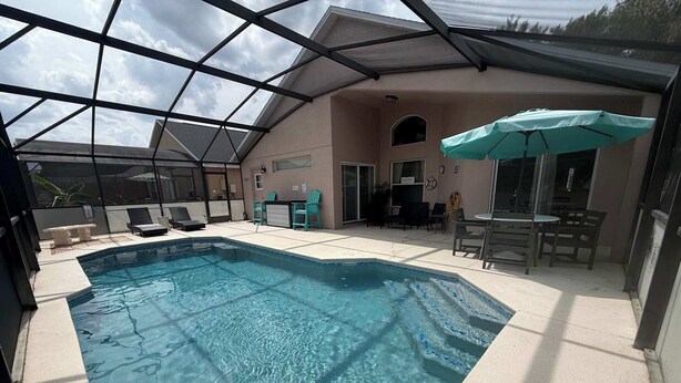 A covered swimming pool with lounge chairs and a table, surrounded by a building with a glass enclosure. - Rebecca’s Villa (Haines City)