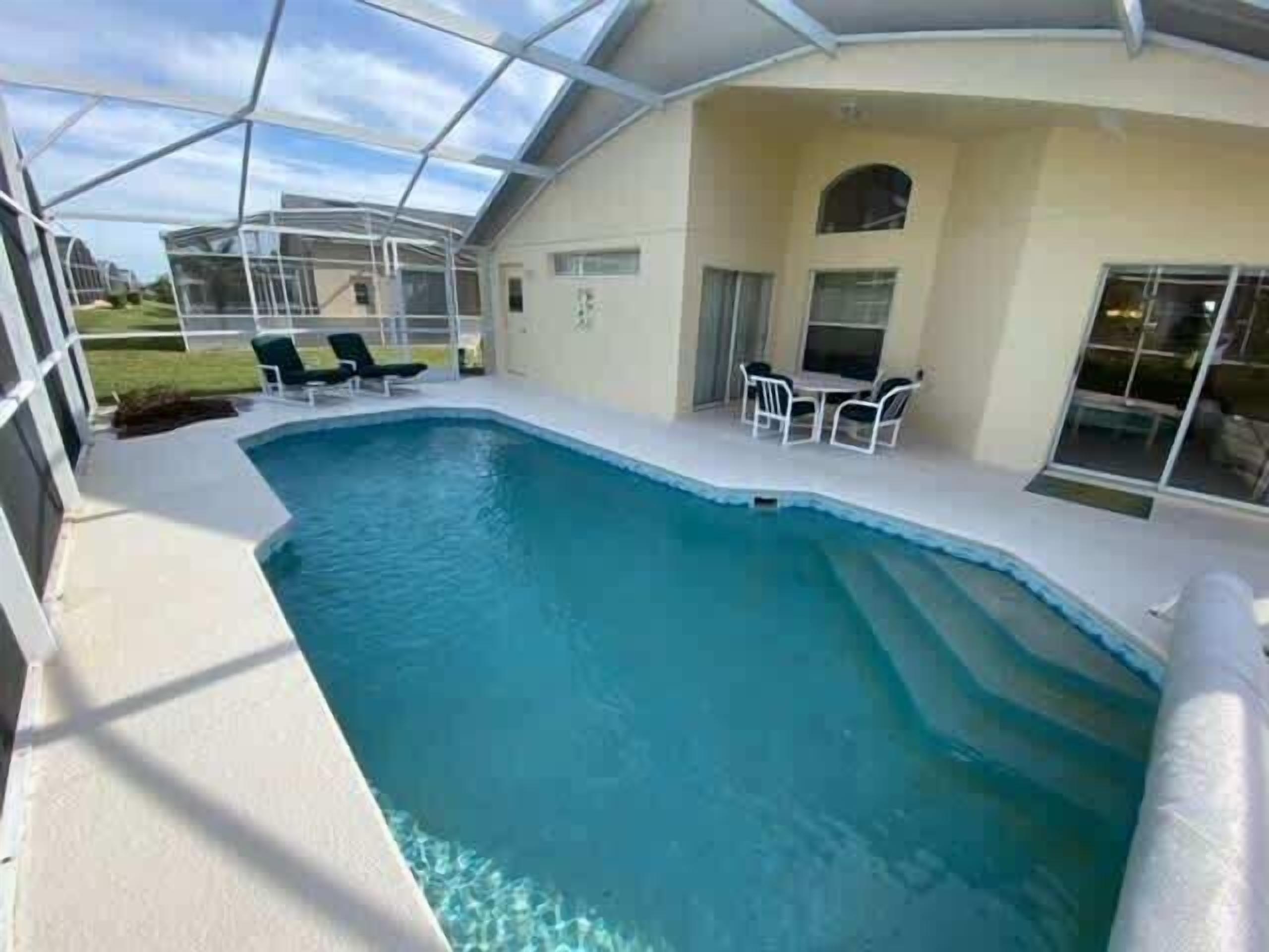 An indoor swimming pool with a glass ceiling, surrounded by a patio area with chairs and a table.