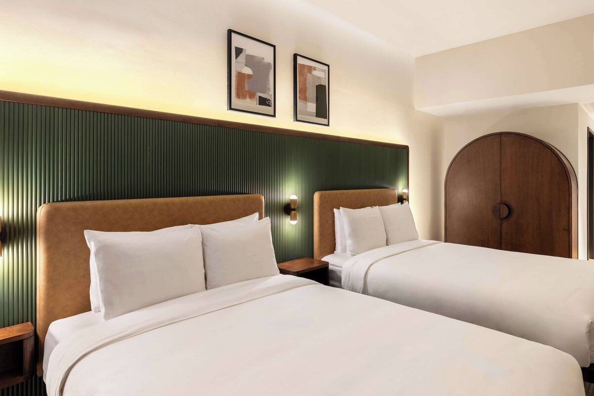 Habitación de hotel con dos camas, cabecero de madera, pared de acento verde y puerta de madera redonda.