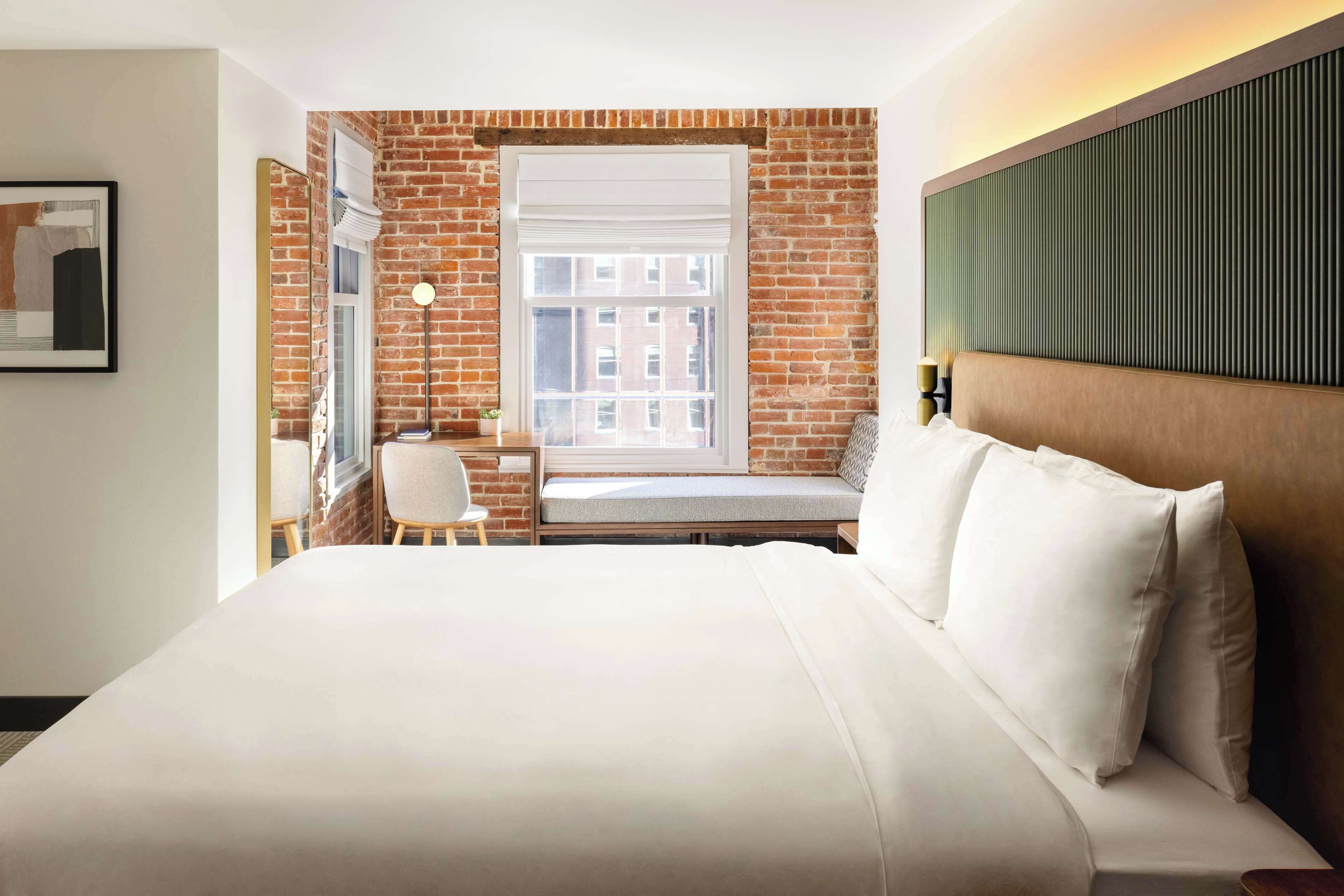Quarto de hotel com uma cama grande, uma escrivaninha com cadeira, uma janela com persianas e uma parede de tijolos.