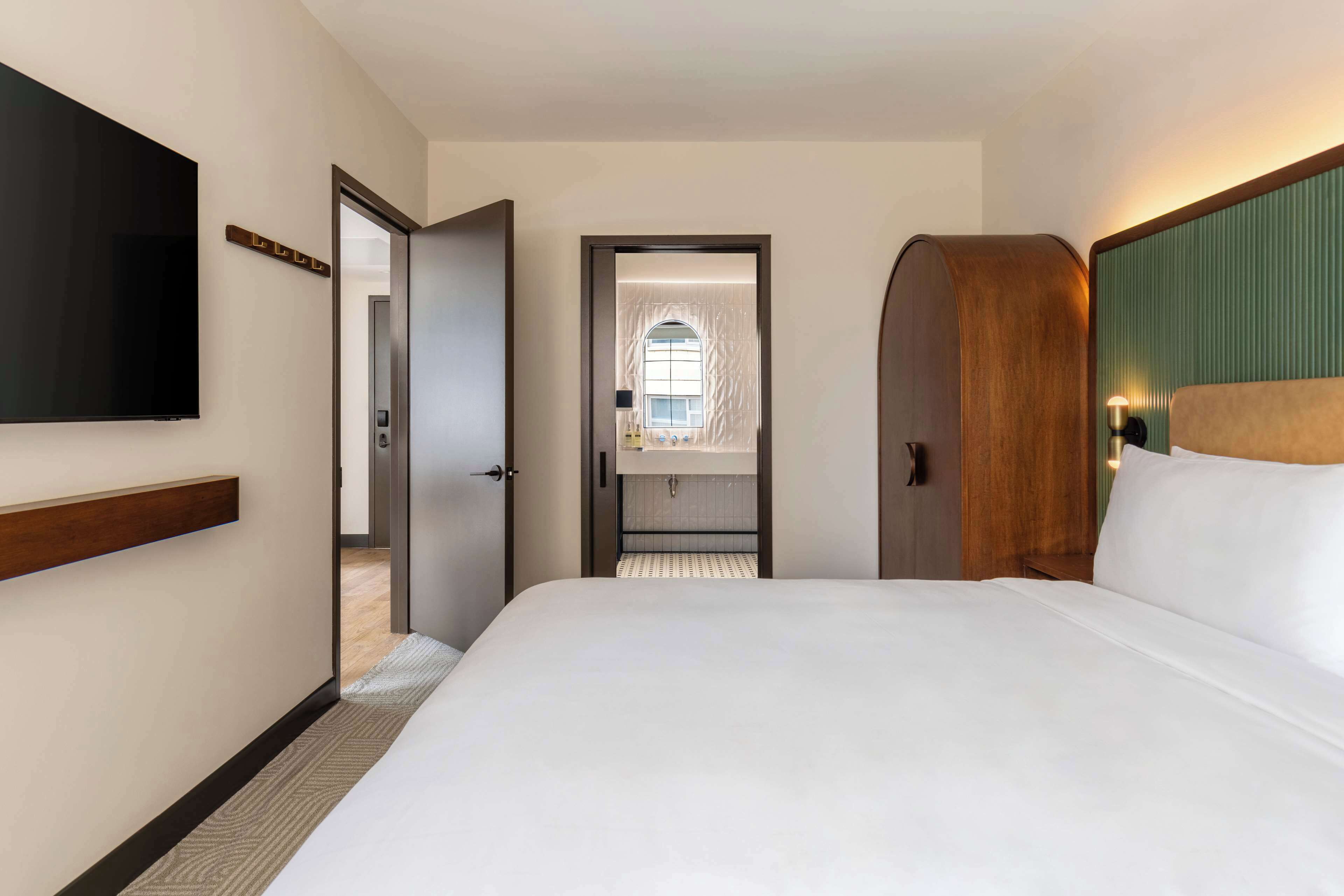 Une chambre d’hôtel dotée d’un grand lit, d’un téléviseur à écran plat fixé au mur, d’une tête de lit en bois et offrant une vue sur le couloir et une autre chambre, grâce à une porte ouverte.