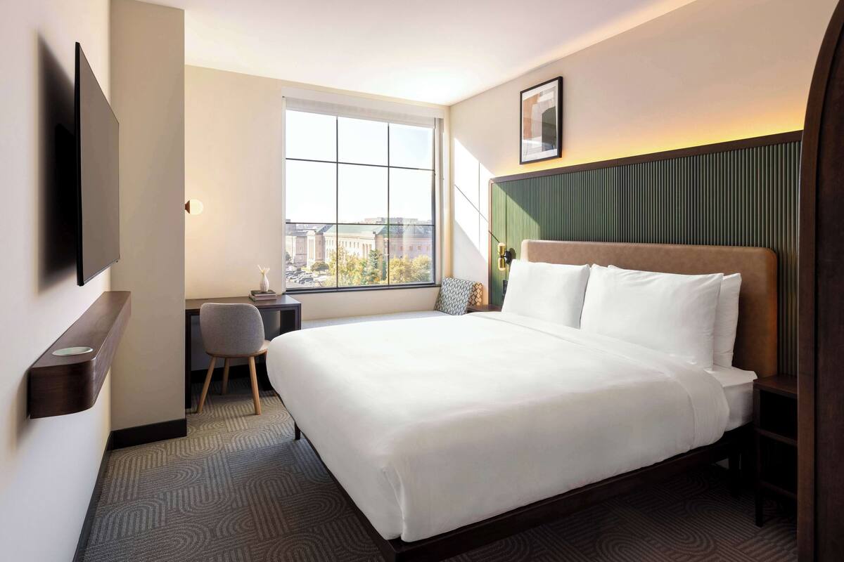 Habitación de hotel con una cama grande, un escritorio, una silla, un televisor montado en la pared y una ventana con vistas a la ciudad.