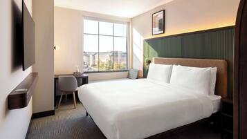 Habitación de hotel con una cama grande, un escritorio, una silla, un televisor montado en la pared y una ventana con vistas a la ciudad.