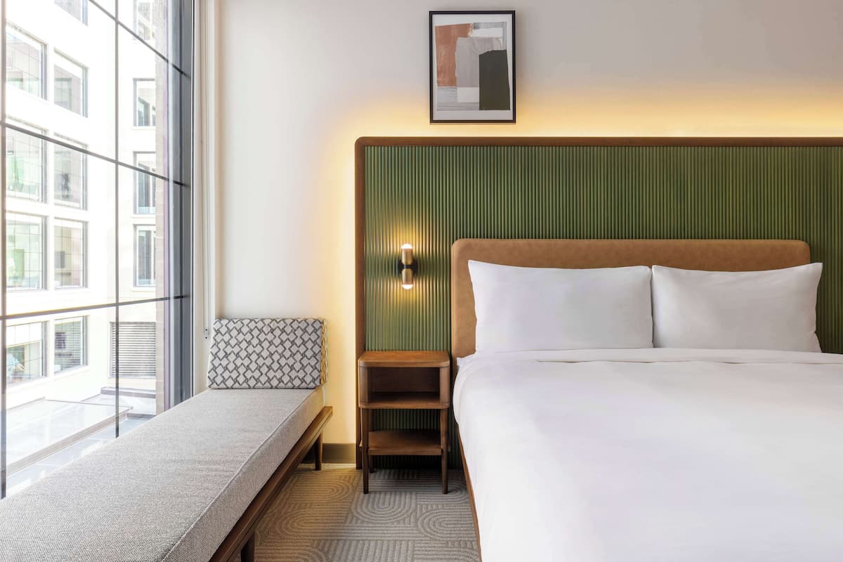 Habitación de hotel con una cama grande, una mesita de noche, un banco y vista a la ciudad a través de un ventanal amplio.