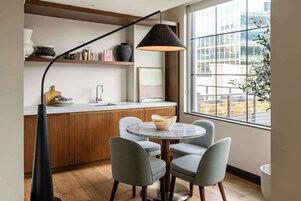 Cozinha moderna com armários de madeira, bancada de mármore e uma mesa de jantar redonda com quatro cadeiras. Uma luminária pendente preta paira sobre a mesa, e uma janela oferece vista da cidade.