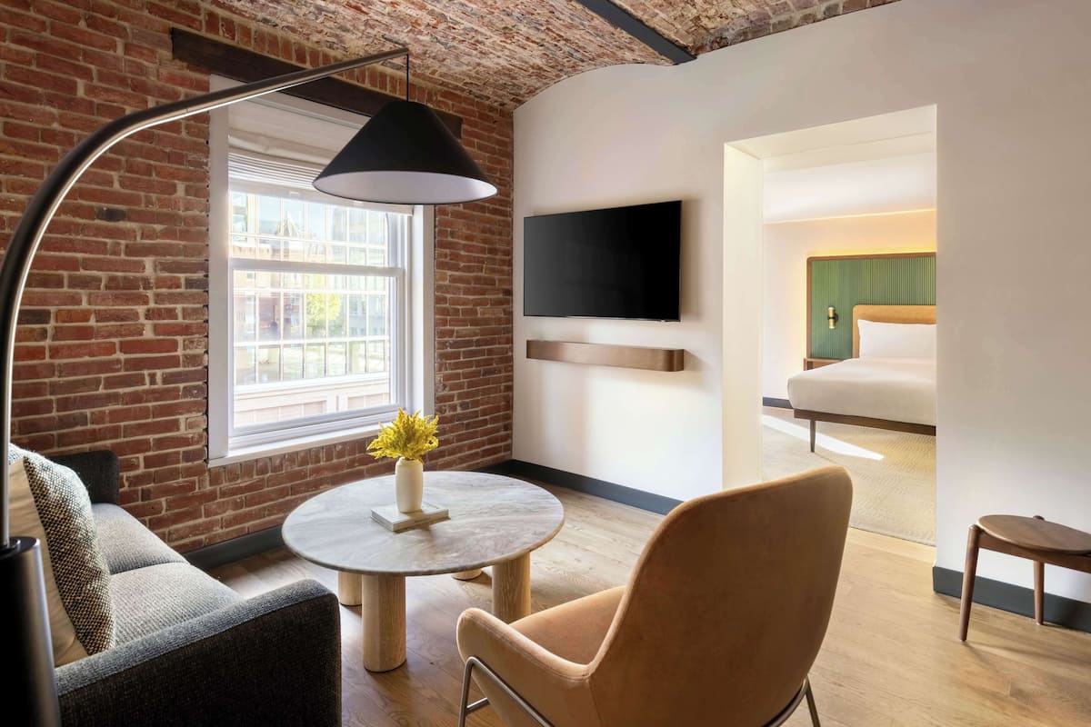Una habitación de hotel moderna con pared de ladrillo, una mesa redonda con un florero, un sofá, un televisor montado en la pared y una cama con cabecera verde.