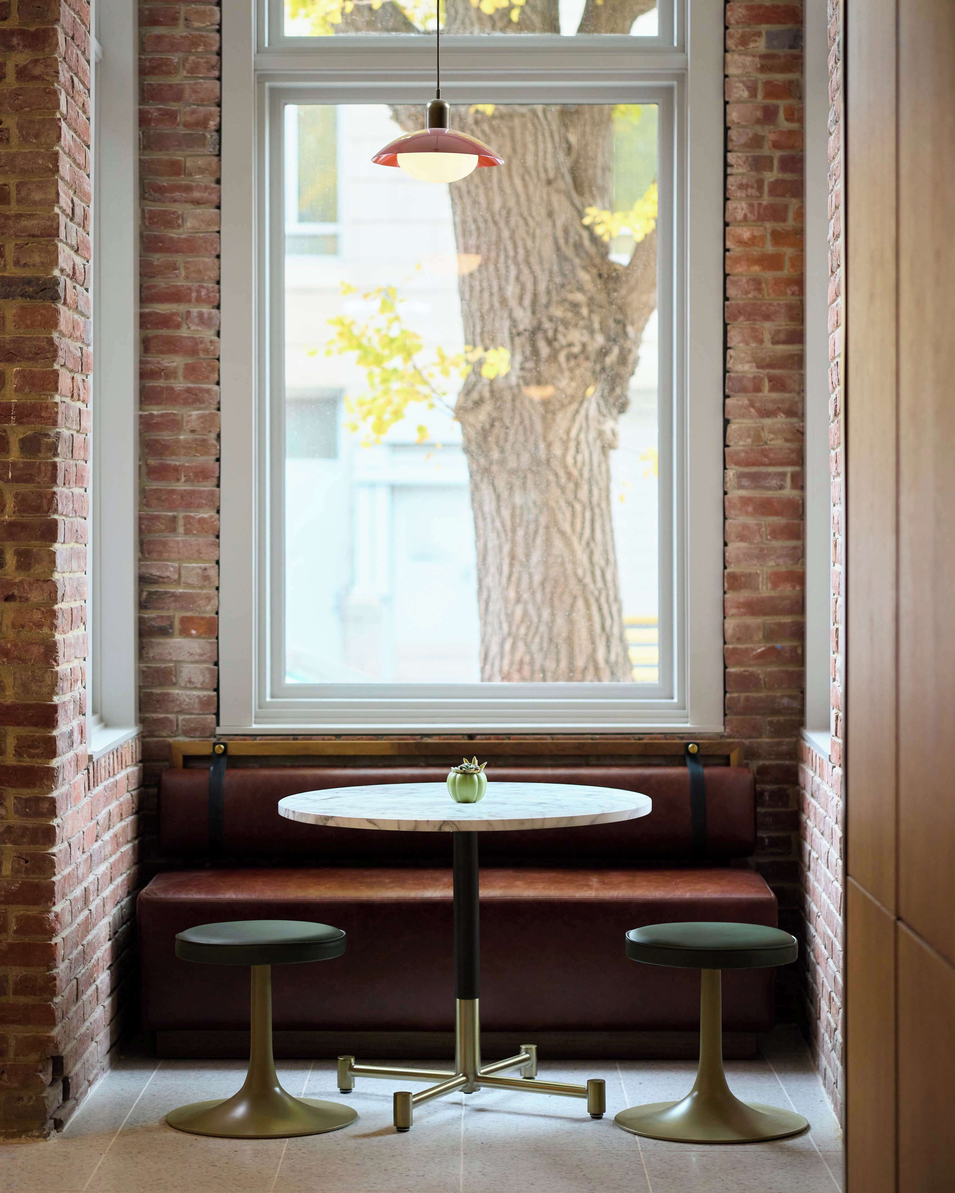 A round table with a green vase, two stools, and a hanging light fixture in front of a window with a brick wall and a tree outside.