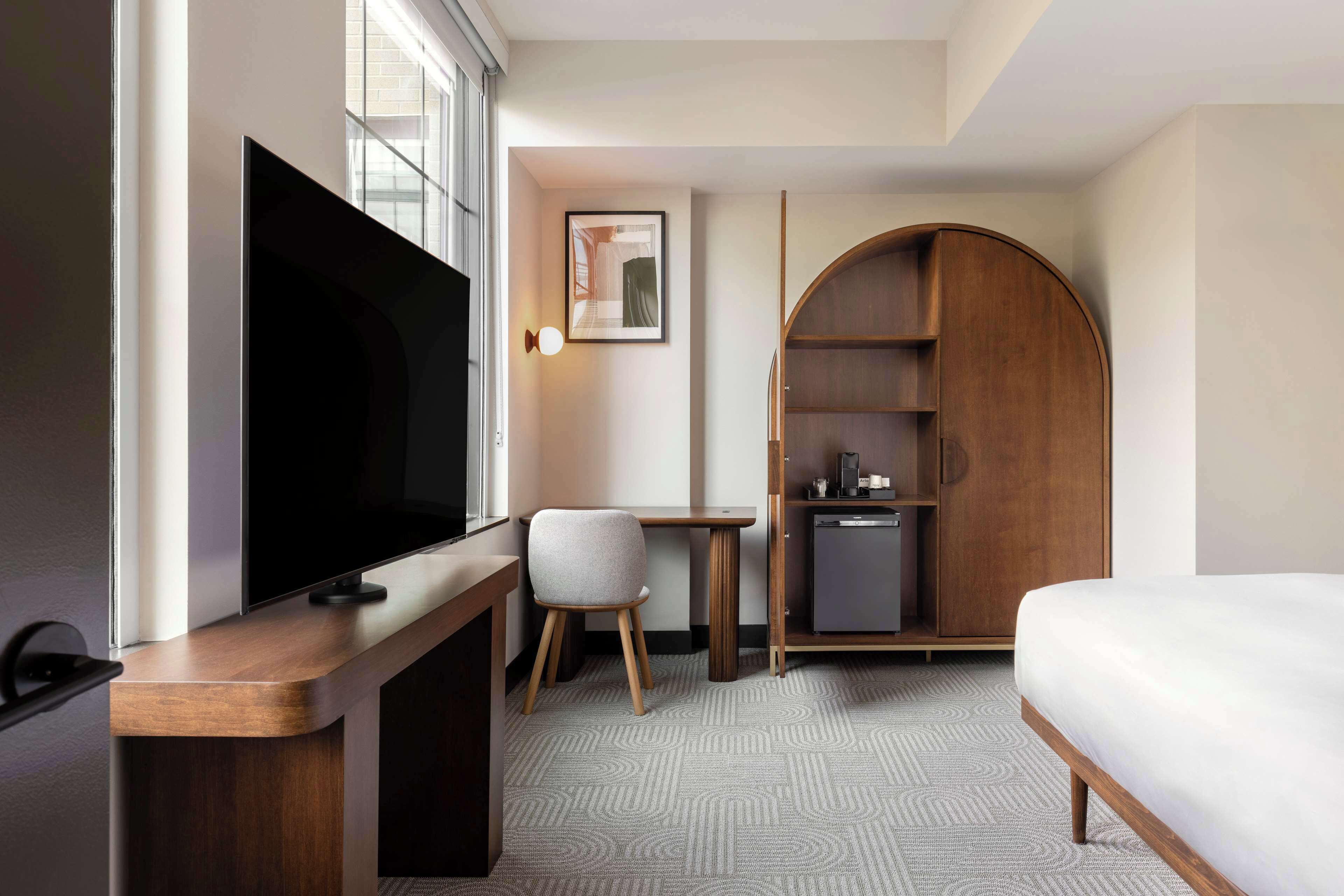 A hotel room with a bed, a desk with a chair, a television, a wardrobe, and a small kitchenette.