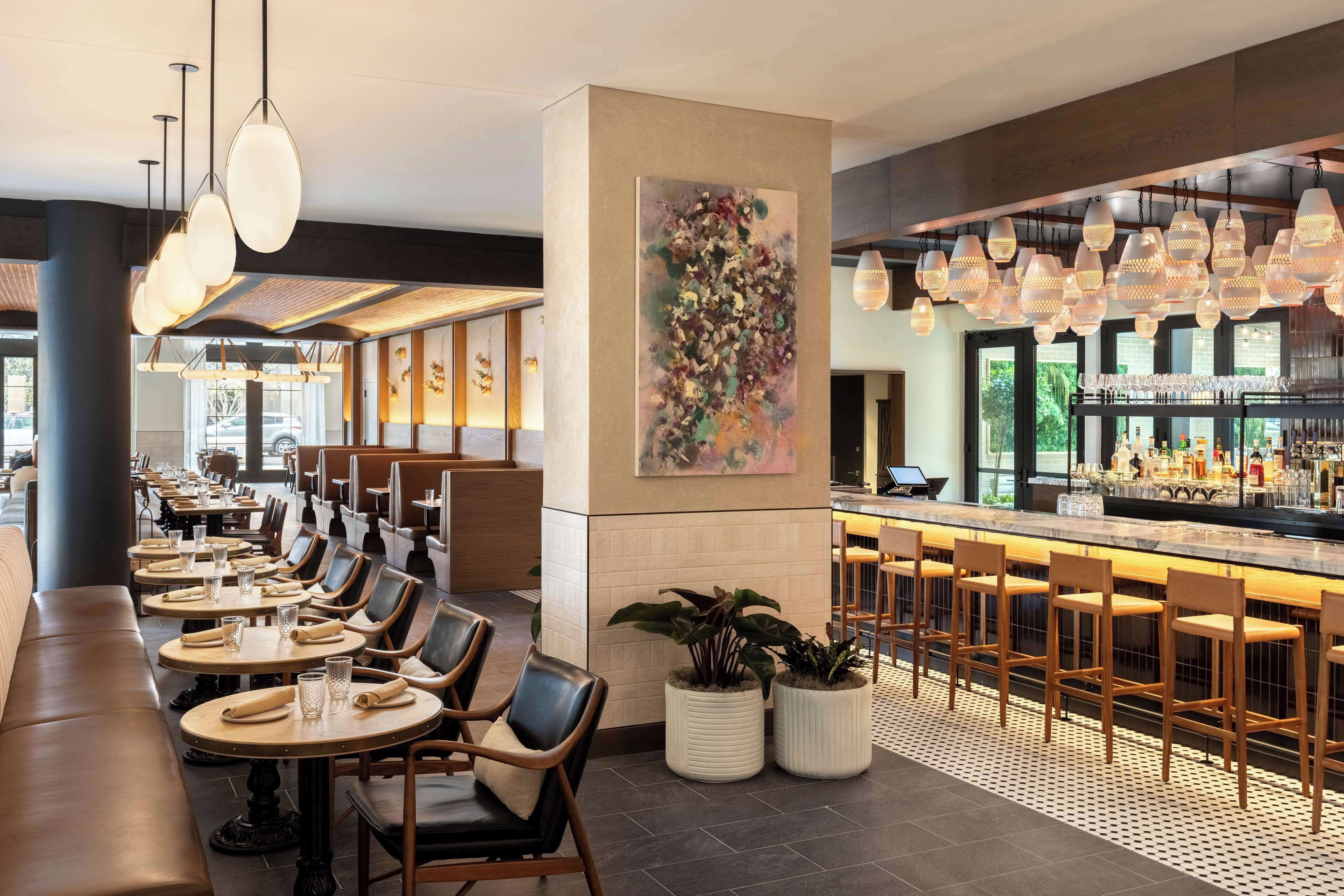 Un restaurant moderne doté de nombreuses tables et chaises, d’un bar avec des tabourets et d’un grand tableau abstrait accroché au mur.