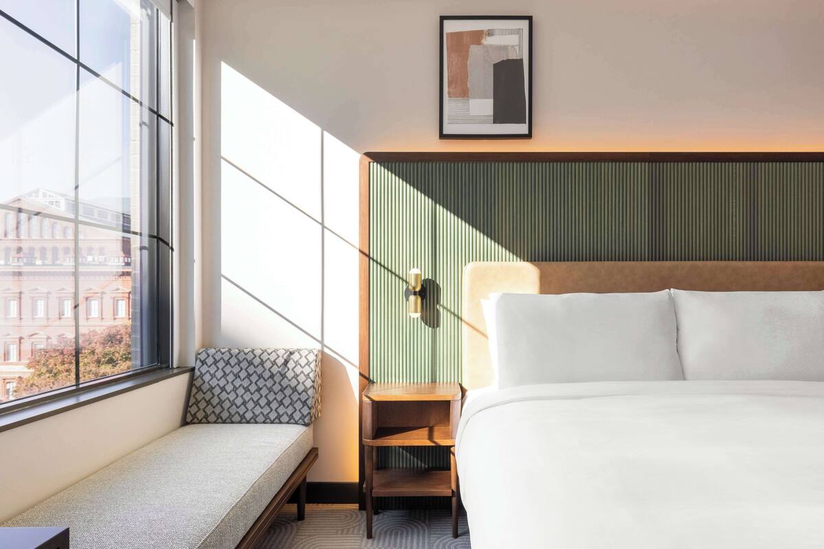 Habitación de hotel con un ventanal grande, una cama, una mesita de noche, una lámpara y una obra de arte colgada en la pared.