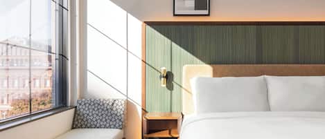 A hotel room with a large window, a bed, a bedside table, a lamp, and a wall-mounted artwork.