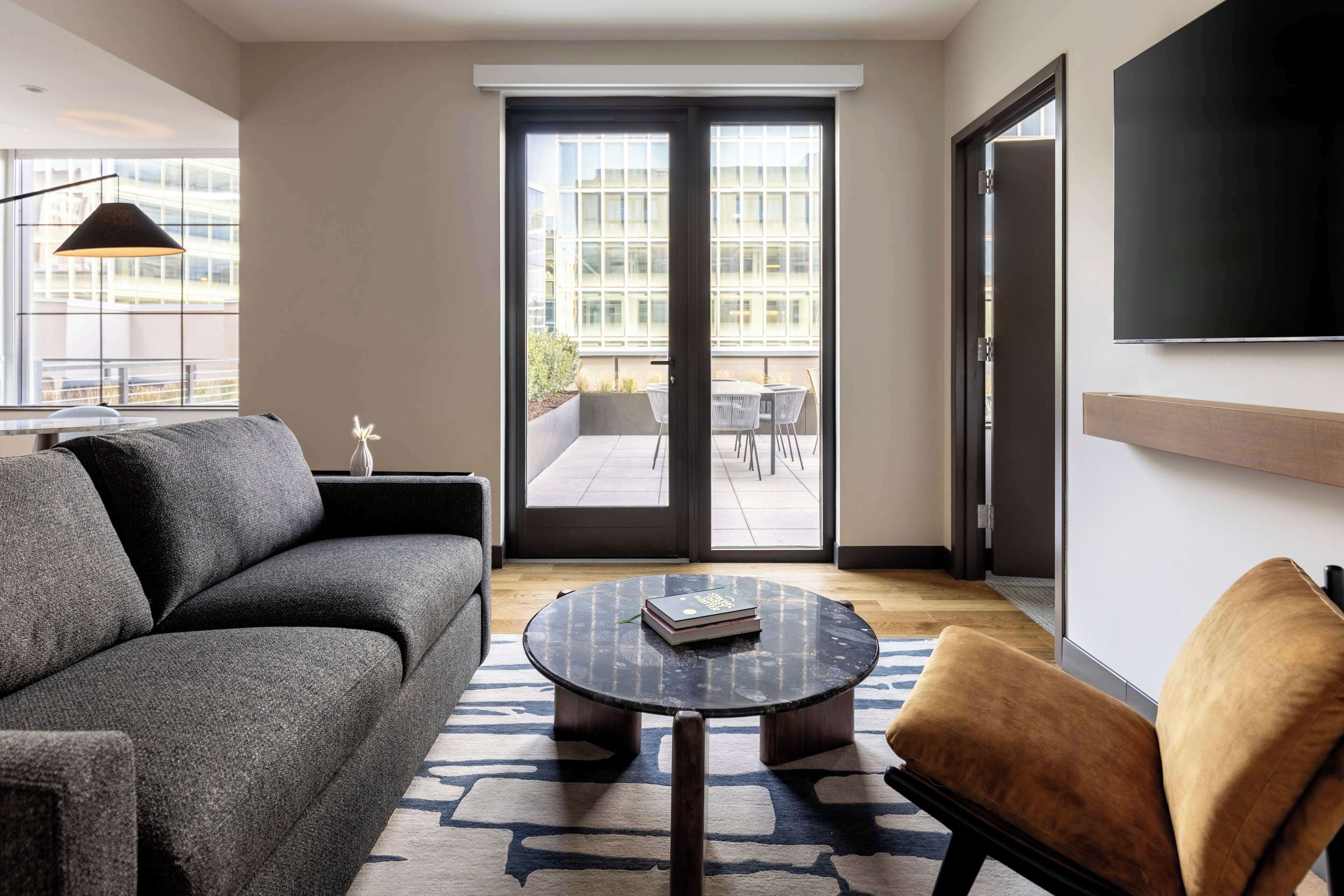 Una sala de estar moderna con un sofá, una mesita redonda y una silla. Hay un ventanal corredero que da a un espacio exterior con una mesa y sillas.