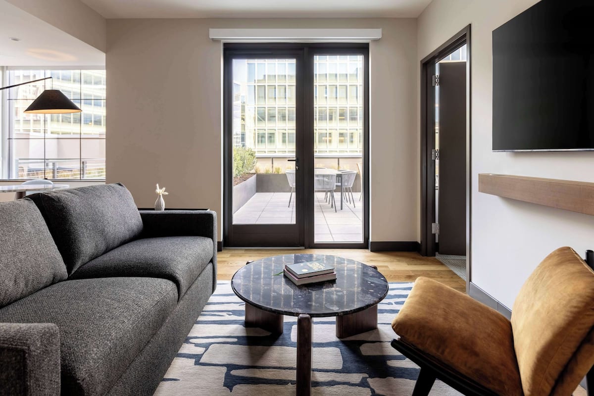 Una sala de estar moderna con un sofá, una mesita redonda y una silla. Hay un ventanal corredero que da a un espacio exterior con una mesa y sillas.