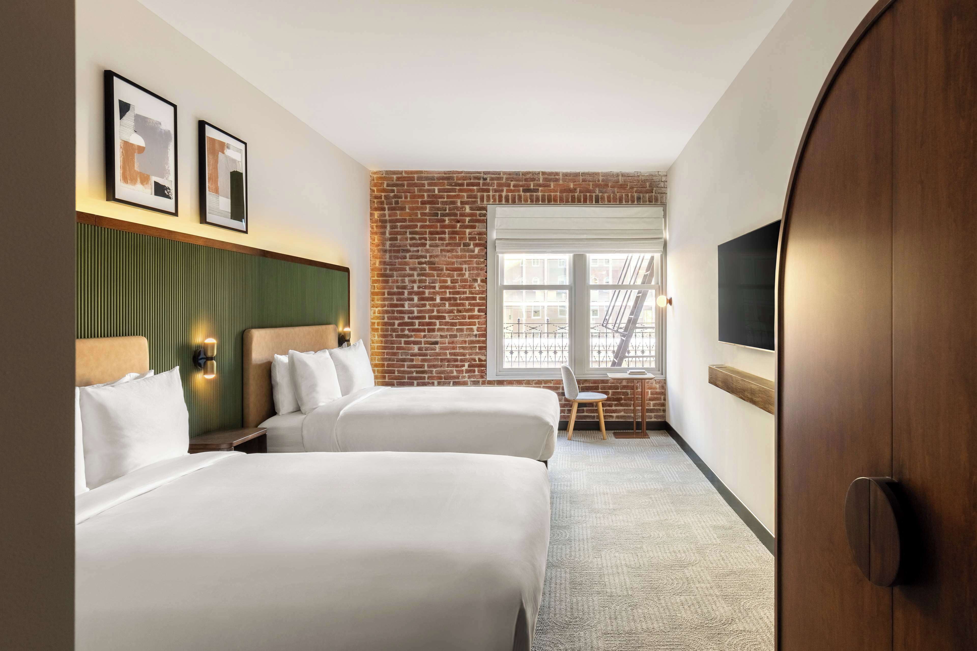 Habitación de hotel con dos camas, una pared de ladrillo, una ventana con vistas y un televisor montado en la pared.