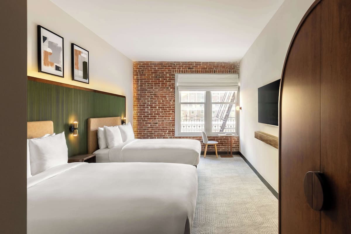 Habitación de hotel con dos camas, una pared de ladrillo, una ventana con vistas y un televisor montado en la pared.