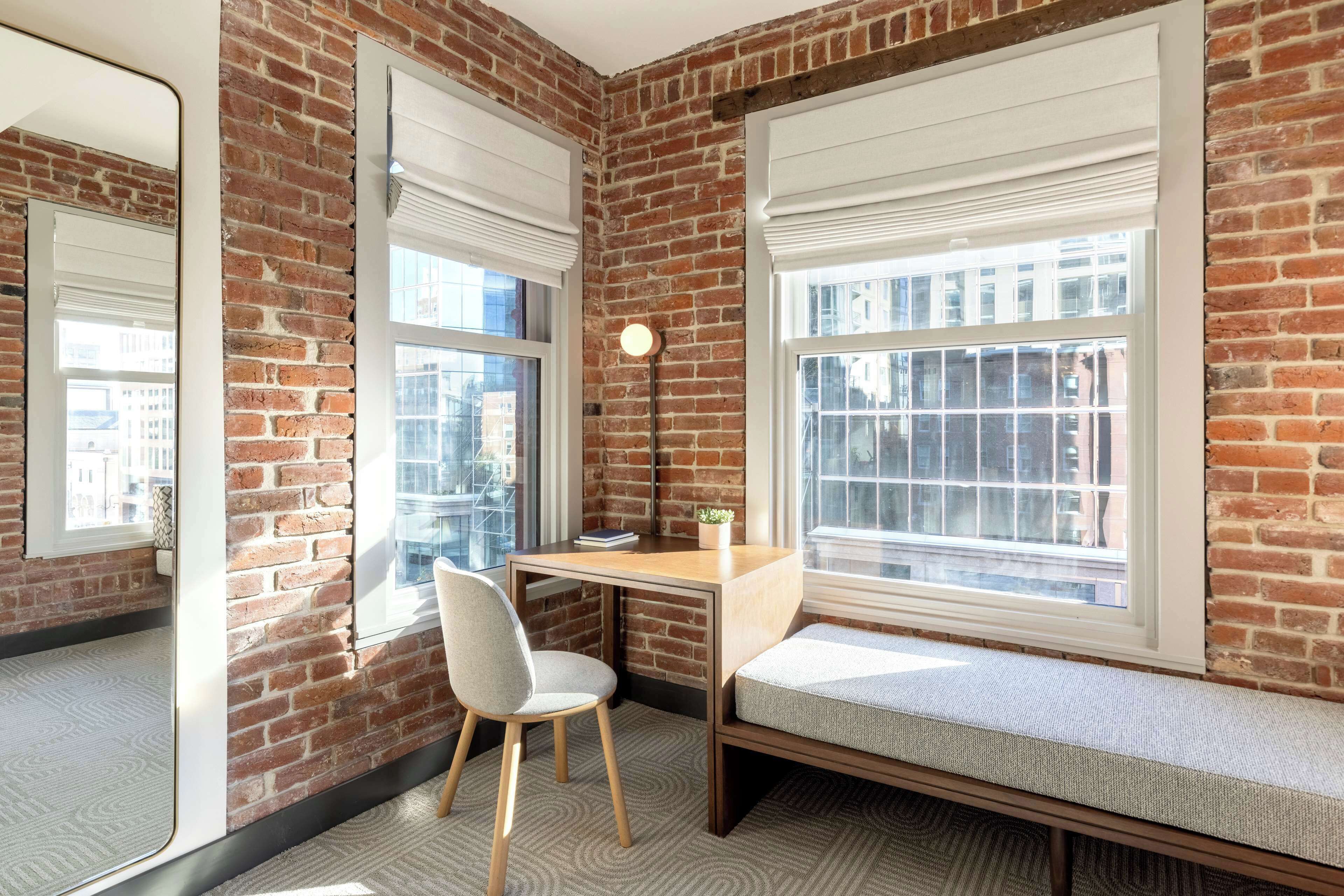 Une pièce avec un mur en briques, un bureau avec une chaise, un banc et de grandes fenêtres.