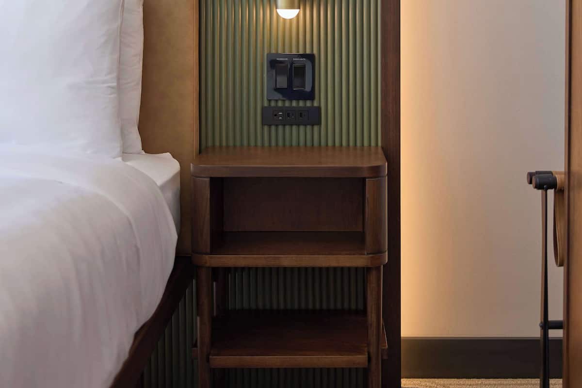 Una habitación de hotel con cama, mesita de noche con lámpara y panel de control en la pared.