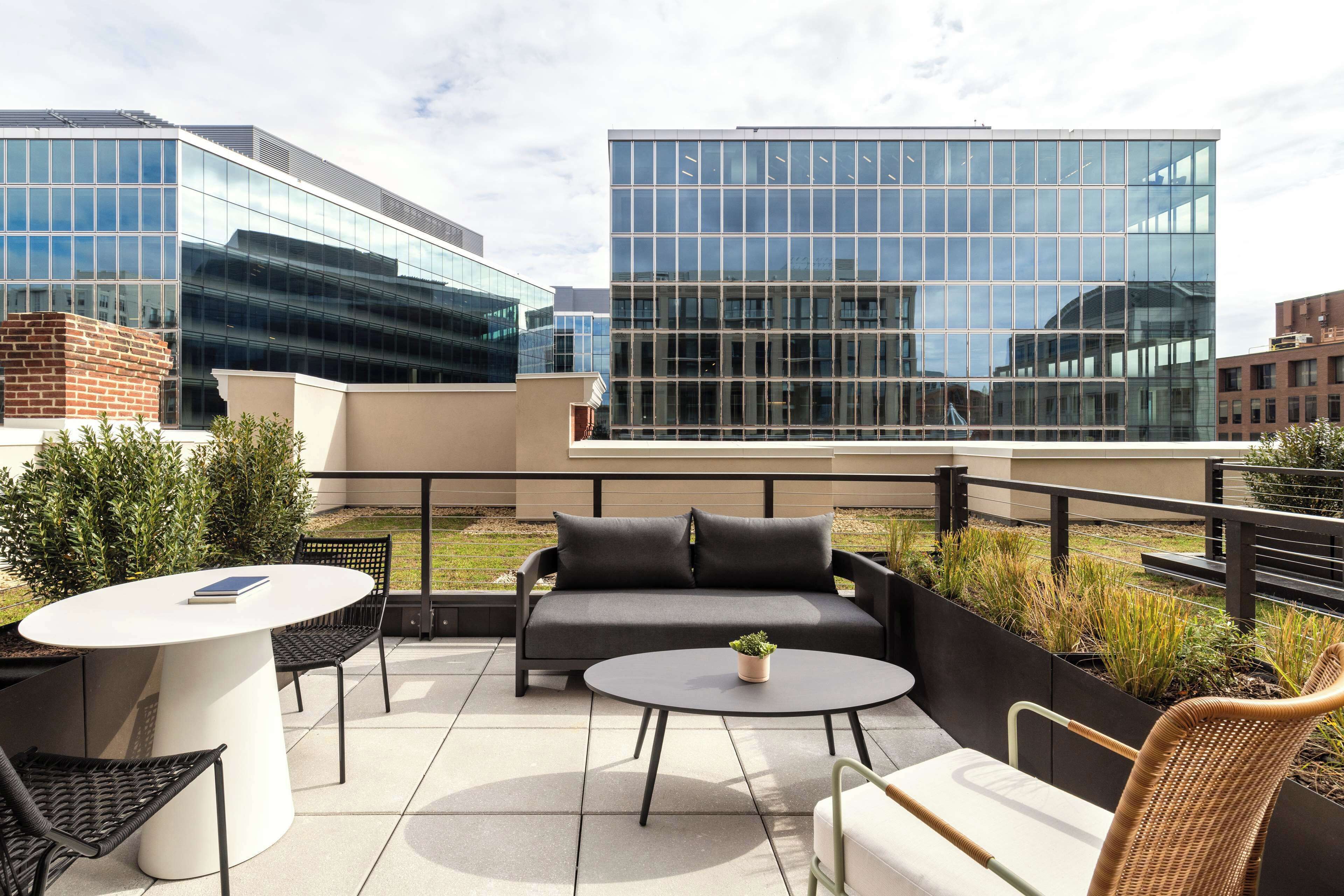 A rooftop terrace with a white round table, black chairs, a grey sofa, and a small potted plant.
