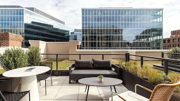 A rooftop terrace with a white round table, black chairs, a grey sofa, and a small potted plant.