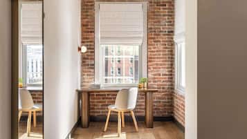 A corner office with a brick wall, a large window with blinds, a desk, a chair, and a small table with a plant.