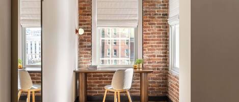 A corner office with a brick wall, a large window with blinds, a desk, a chair, and a small table with a plant.