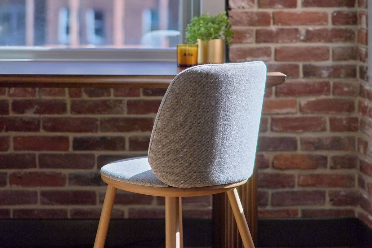Una silla con asiento de tela texturizada y patas de madera, ubicada frente a una ventana con vista a un edificio de ladrillo.