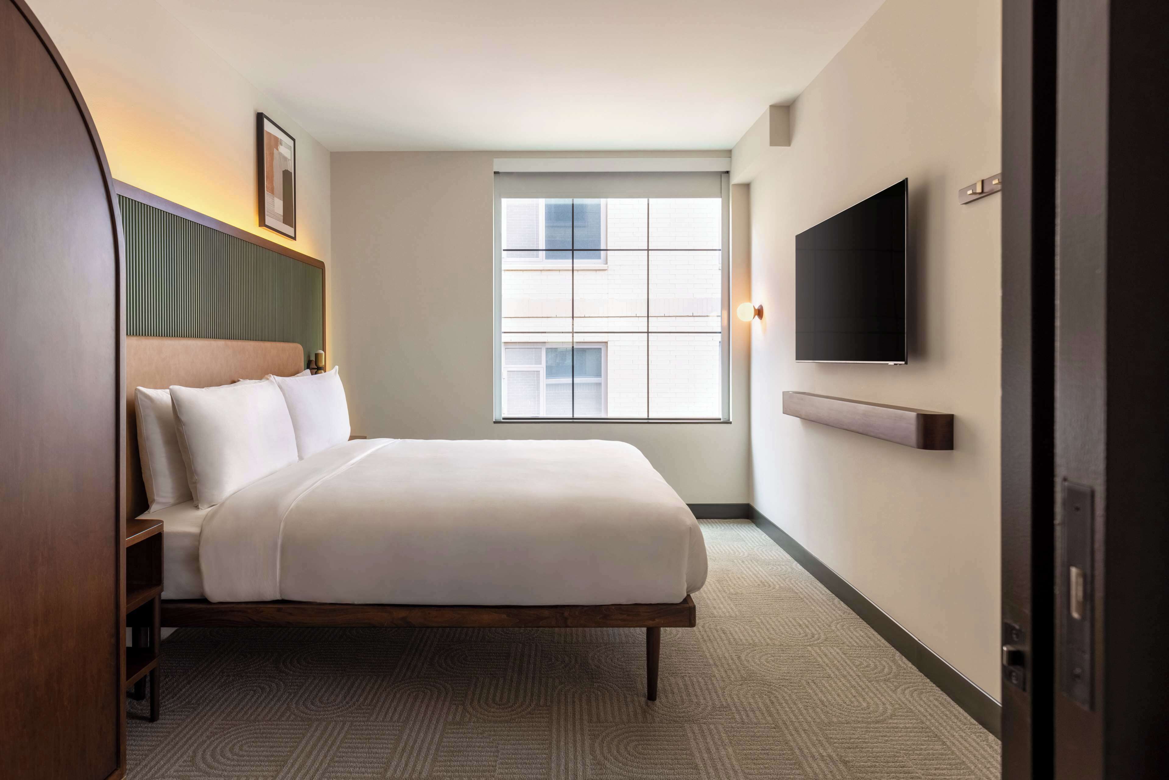 A hotel room with a bed, a TV mounted on the wall, a window with blinds, and a door.