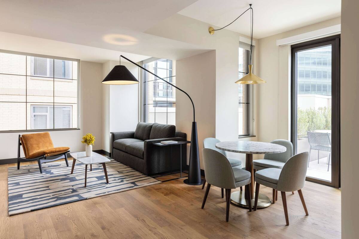 Una sala de estar moderna con un sofá, una mesa de centro y un comedor con sillas. Hay ventanas amplias que proporcionan luz natural y vistas al exterior.