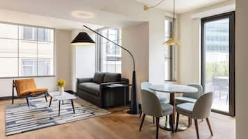 A modern living room with a sofa, a coffee table, and a dining area with chairs. There are large windows providing natural light and a view of the outside.