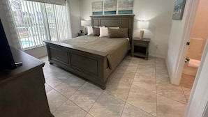 A bedroom with a large bed, a ceiling fan, a nightstand with a lamp, and a view of the outdoors through the window. - Azure Palms Retreat (Haines City)