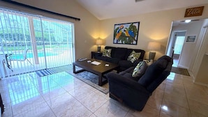 A spacious living room with a large sliding glass door leading to a patio, a black sofa set, a coffee table, and a painting of parrots on the wall. - Prestige on the Fairway (Haines City)