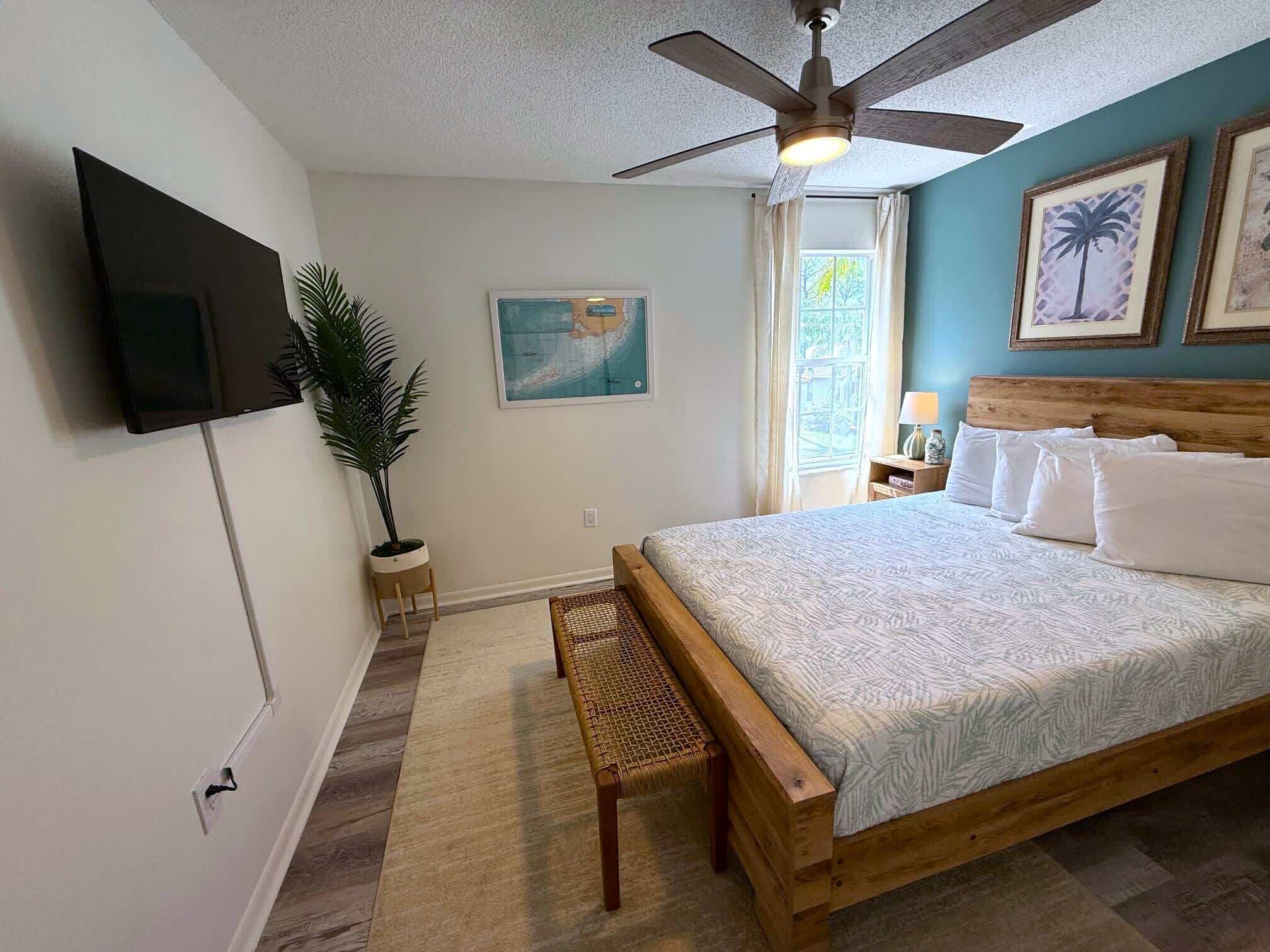 Une chambre à coucher comprenant un lit, un ventilateur de plafond, un téléviseur fixé au mur, une fenêtre avec des rideaux et une plante.