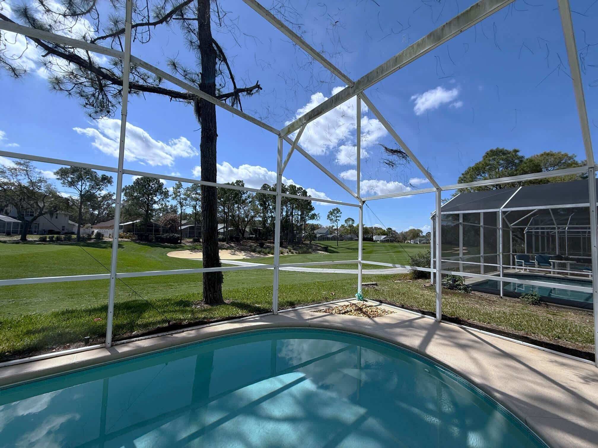 A swimming pool with a covered area, surrounded by a metal fence, with a view of a golf course and houses in the background.