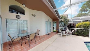 A covered patio with a glass table and chairs, a pool, and a view of greenery. - Oasis of Southern Dunes (Haines City)