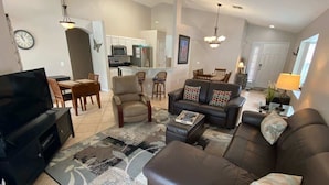 A modern living room with a leather sofa, a coffee table, and a TV. There is a dining area with chairs and a table, and a kitchen in the background. - Oasis of Southern Dunes (Haines City)