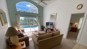 En stue med en sofa, et sofabord og en lampe. Der er et vindue med gardiner og udsigt til en pool.
