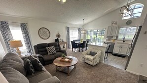 A spacious living room with a grey sofa, a round coffee table, and a patterned armchair. A dining area with a table and chairs is visible in the background. - Stay Awhile at the Fairways (Haines City)